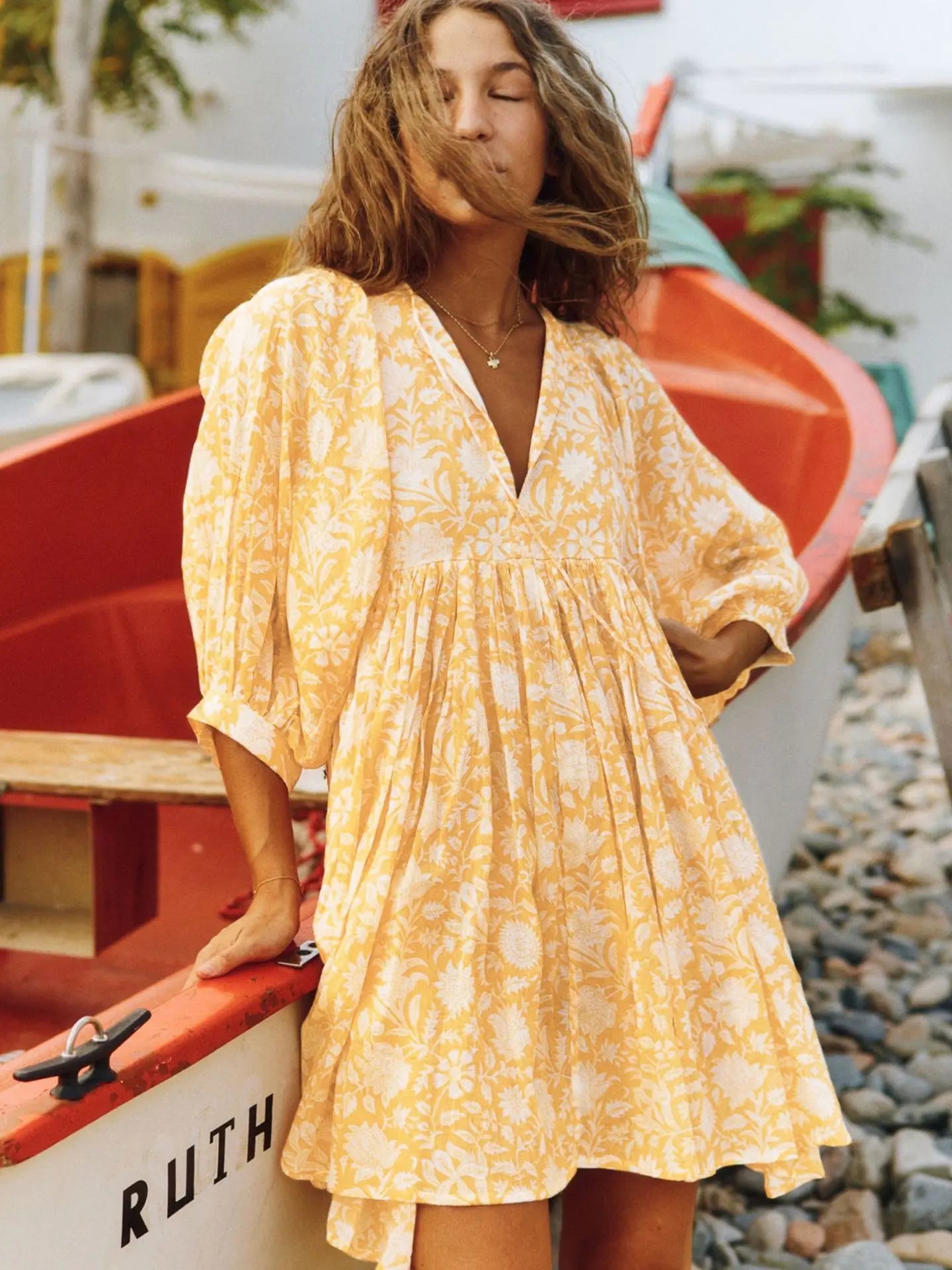 Vestido floral amarelo curto com mangas volumosas e decote em V, em cenário litorâneo com styling descontraído de verão.