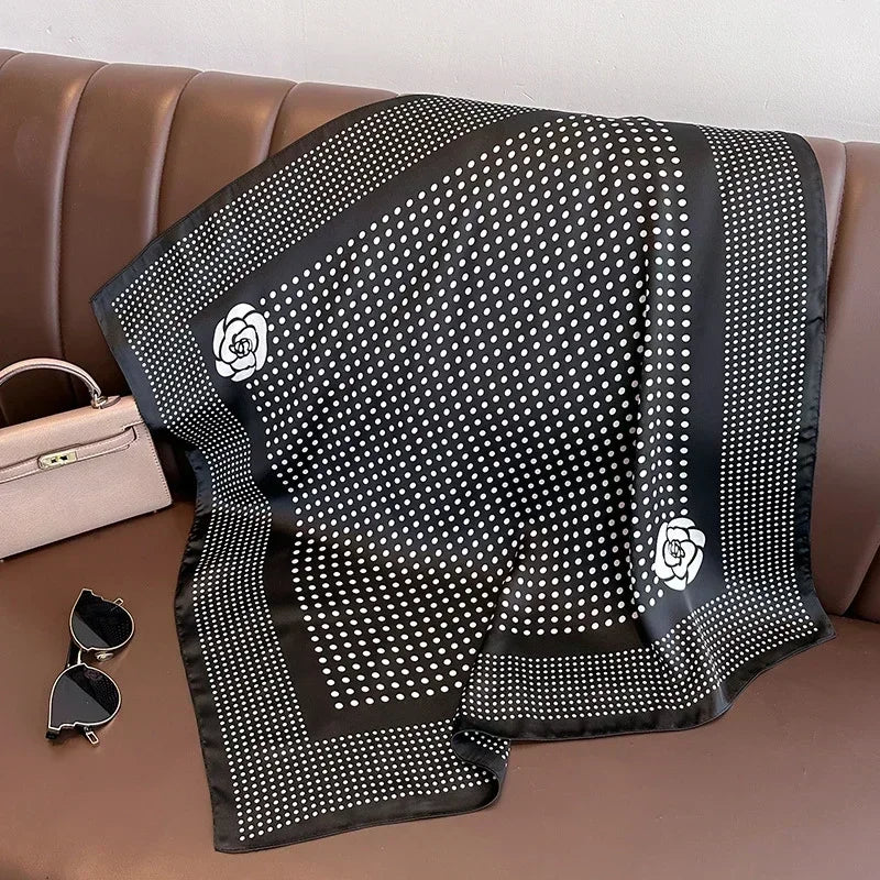 Lenco Versatile Grace em fundo preto com estampa de poa branca e flores minimalistas nas bordas, disposto sobre banco marrom ao lado de bolsa bege e oculos escuros