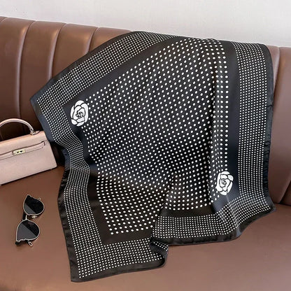 Lenco Versatile Grace em fundo preto com estampa de poa branca e flores minimalistas nas bordas, disposto sobre banco marrom ao lado de bolsa bege e oculos escuros