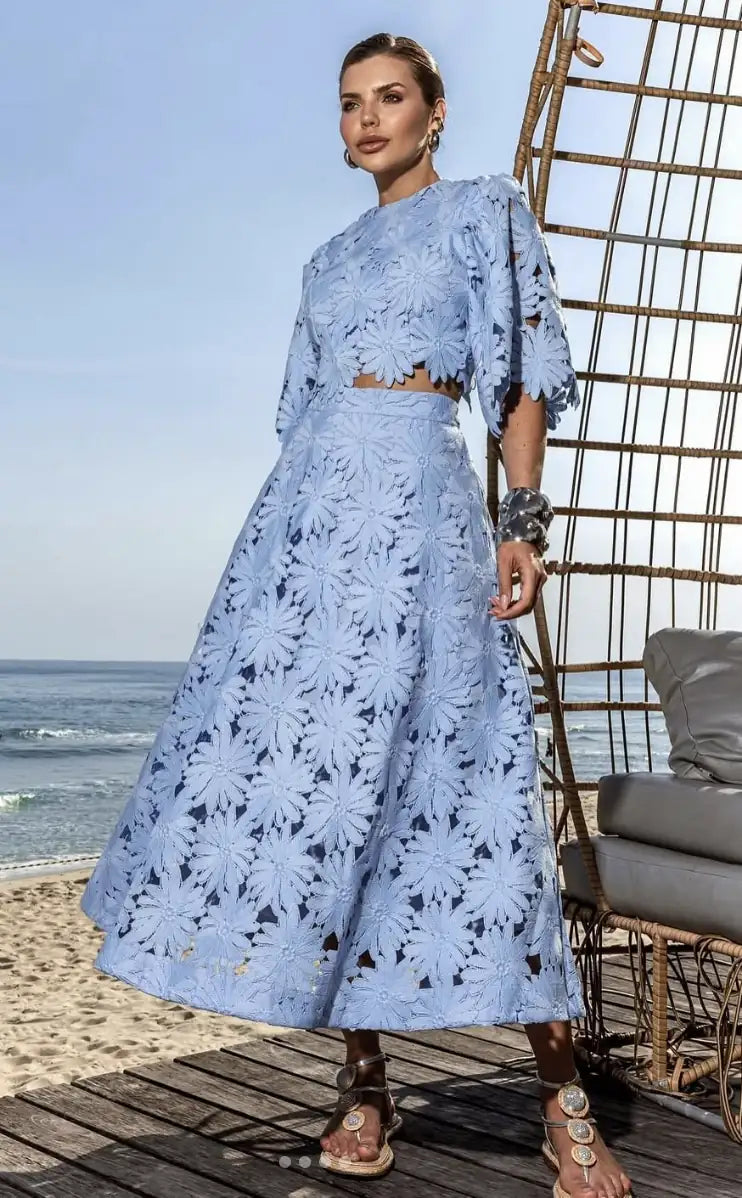 Conjunto Antonella de renda floral azul claro, exibido em look casual e elegante para o dia a dia. A imagem destaca a modelagem da blusa cropped e da saia longa, que valoriza a silhueta em um ambiente externo.