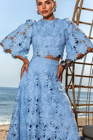 Modelo em conjunto Antonella azul claro com blusa cropped e saia longa A-line, em renda floral. O look, ideal para eventos de verão e praia, é fotografado em um cenário à beira-mar, destacando o caimento da peça.