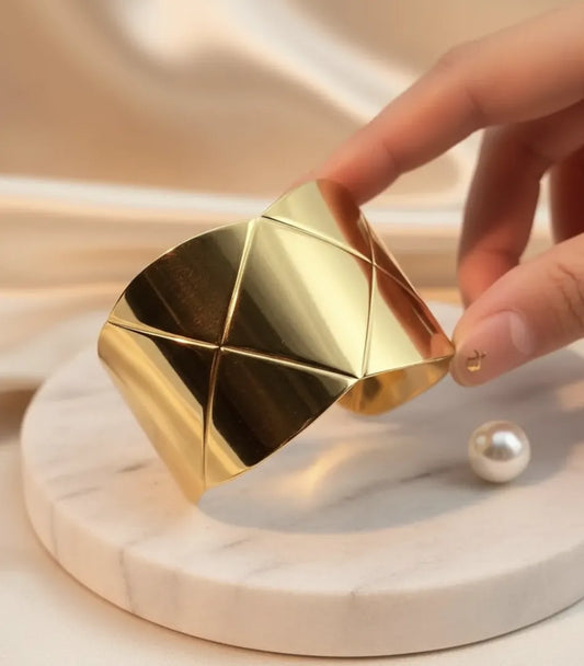 Pulseira dourada da Dress Eleven com textura geométrica e acabamento em aço inoxidável 18K sobre base de mármore claro.