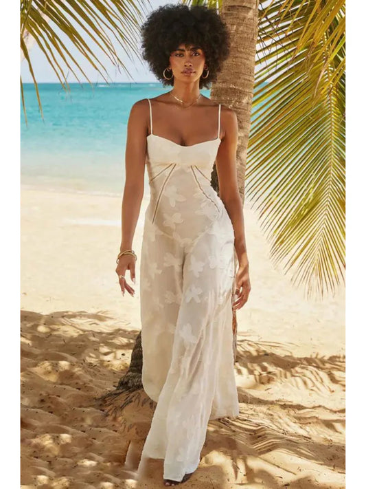 Vestido longo Aveline com alças finas e bordados florais, modelo posando à beira-mar com palmeira ao fundo.