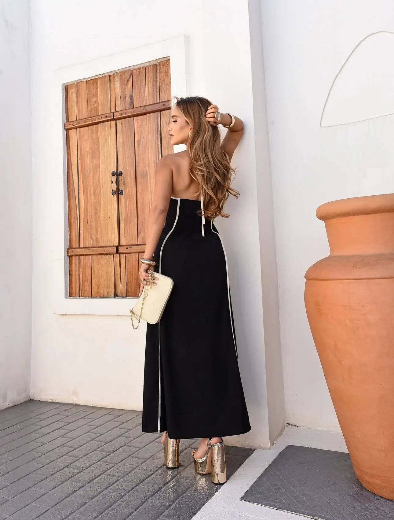 Vestido Dianna preto longo frente única visto de costas, com detalhe de amarração e caimento elegante, modelo segurando clutch clara.