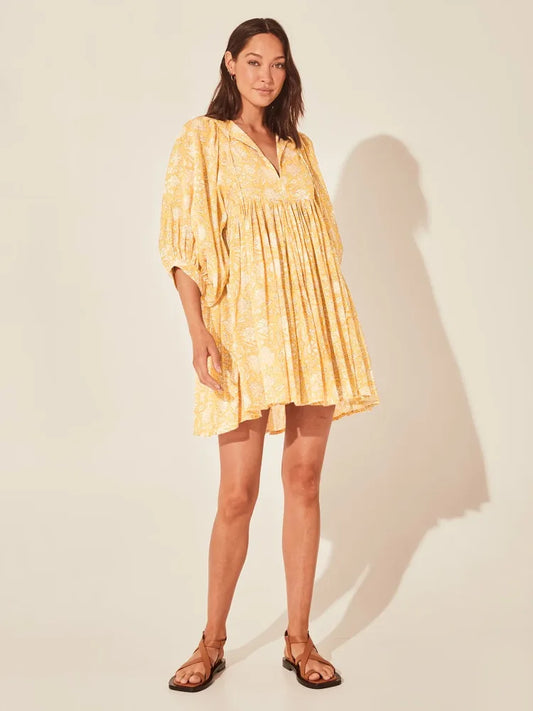 Vestido curto com estampa floral amarela, mangas bufantes e caimento solto, fotografado de frente em estúdio com fundo bege.

