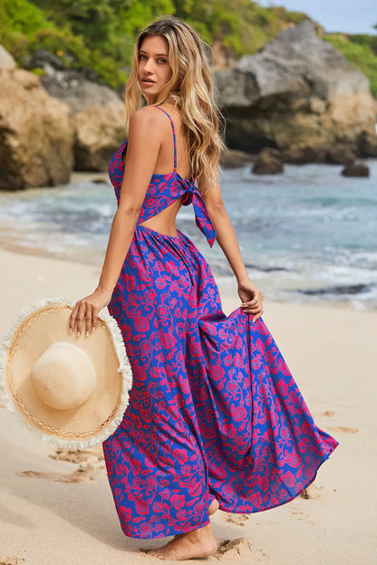 Vestido longo roxo com estampa floral, modelo caminha na praia com chapéu na mão, ideal para verão.