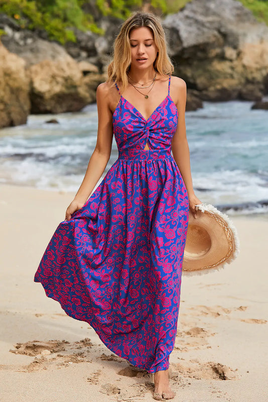 Vestido longo roxo com estampa floral, modelo caminha na praia com chapéu na mão, ideal para verão.

