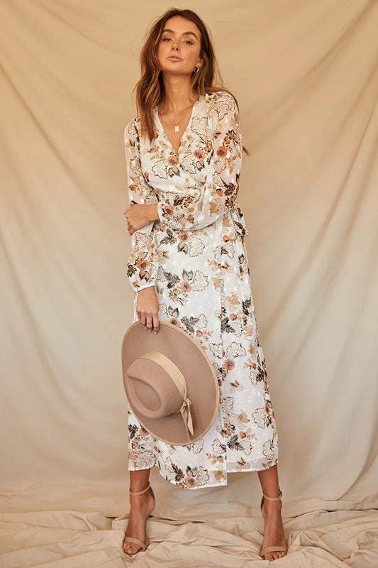 Modelo de vestido Positano longo floral com decote V e manga comprida, segurando chapéu de palha em ensaio fotográfico de estúdio, ideal para eventos diurnos.
