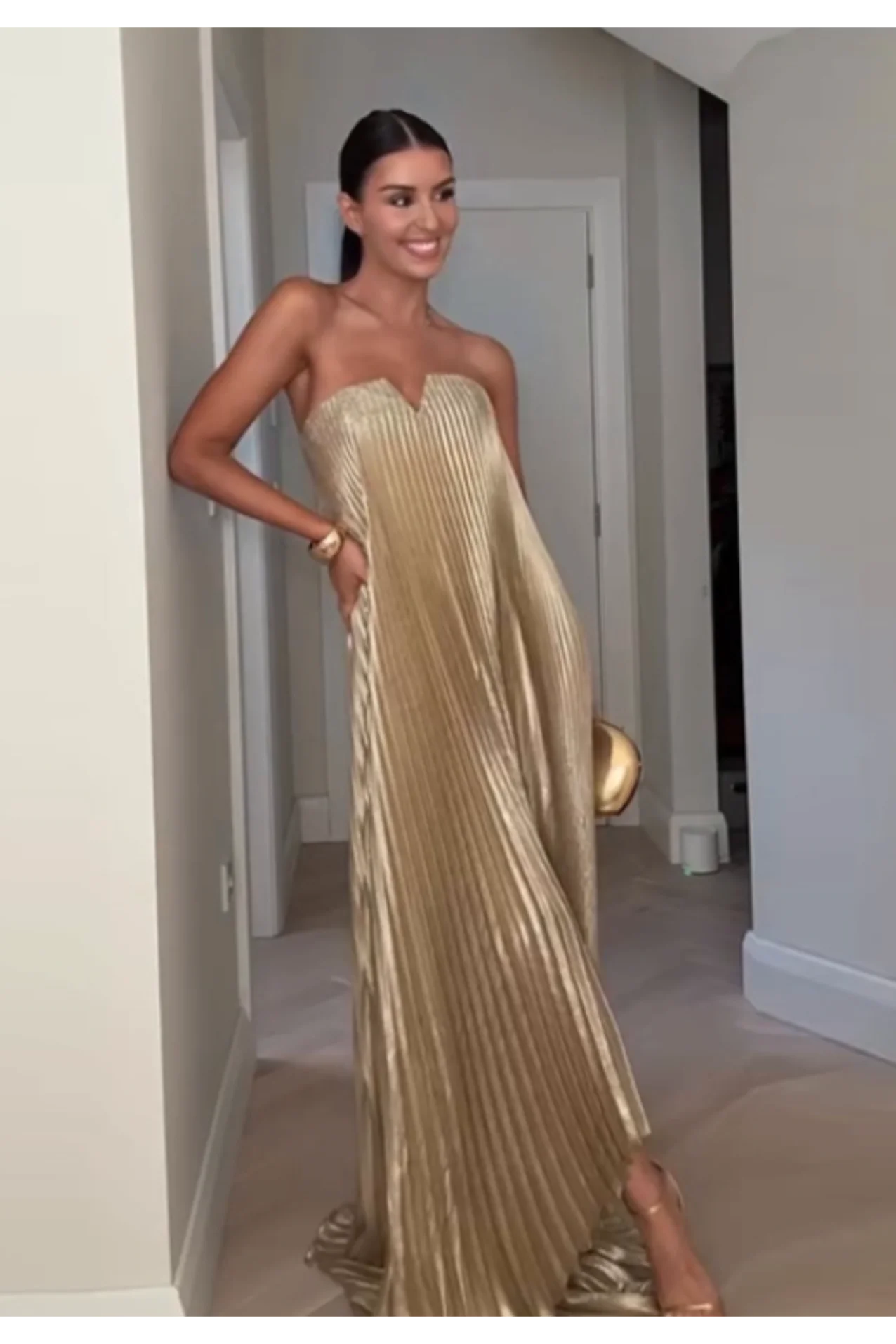 Mulher usando vestido plissado dourado com alças finas e clutch metálica em ambiente interno, vestido ideal para festas elegantes