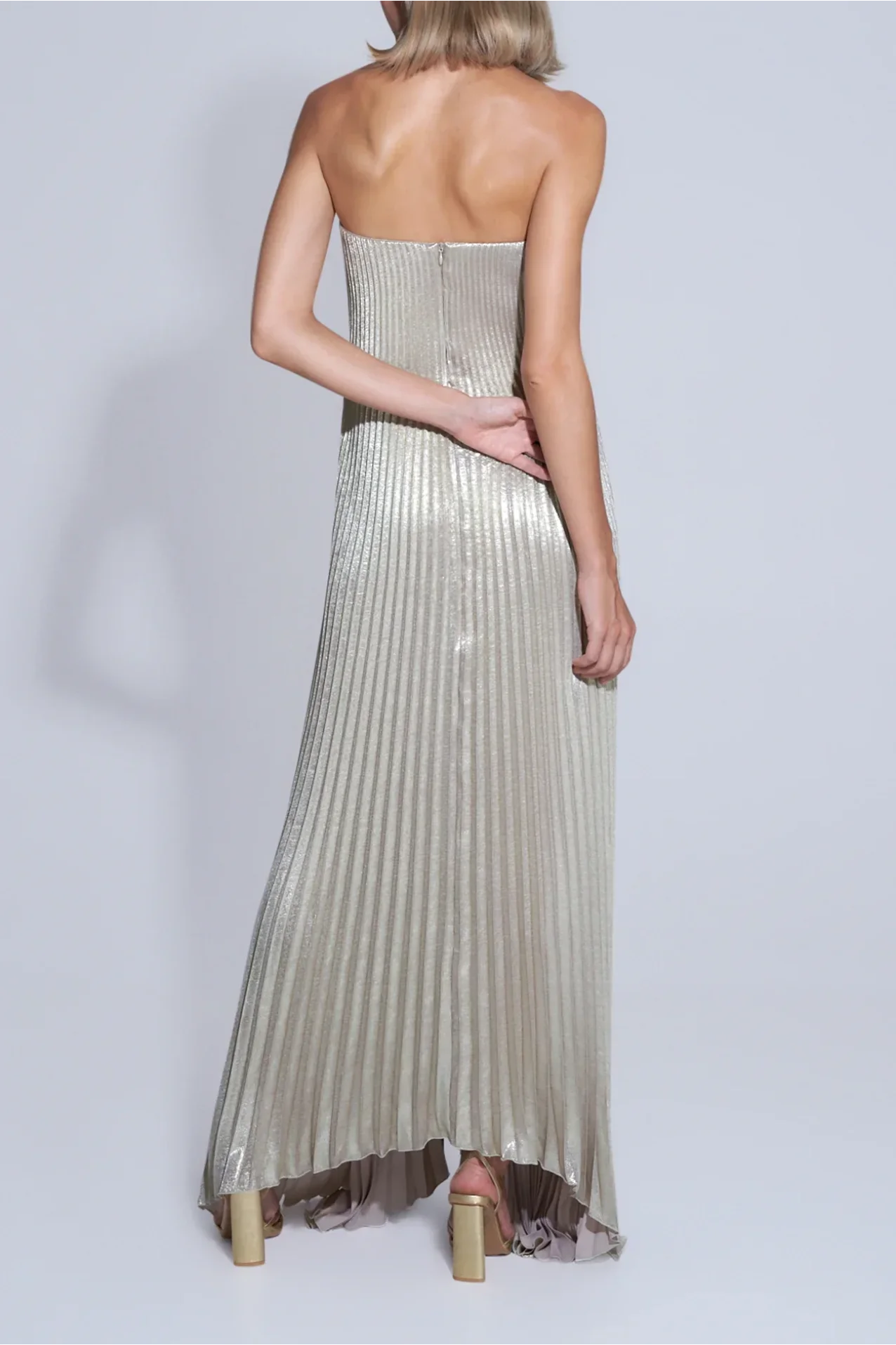 Costas do vestido plissado dourado com decote reto sem alças, modelo loira de perfil em fundo claro